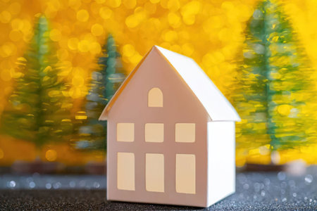 Small white paper house model on a dark surface with blurred christmas trees and festive lights in...