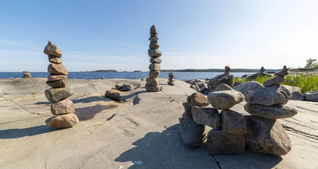 Stacks of stones are balanced on a rocky shoreline overlooking the ocean on a clear sunny day with a...