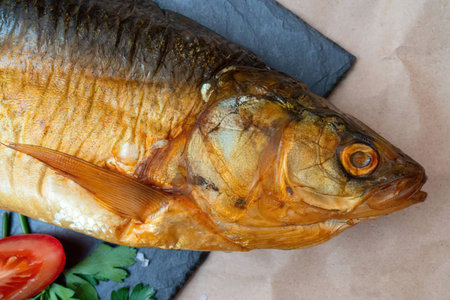 Smoked fish lies on a black slate serving board accompanied by fresh herbs and a tomato slice.