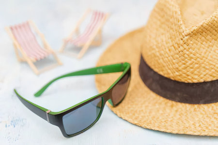 Green sunglasses and a straw hat with a brown band lie on a beach, ready for a relaxing summer...