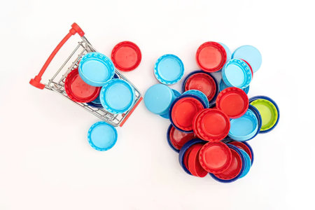 Colorful plastic bottle caps spilling out from a miniature shopping cart onto a plain white surface,...