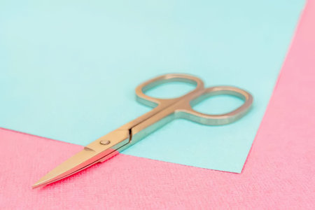 Gold scissors with sharp pointed tips resting on pastel blue and pink paper background symbolizing...