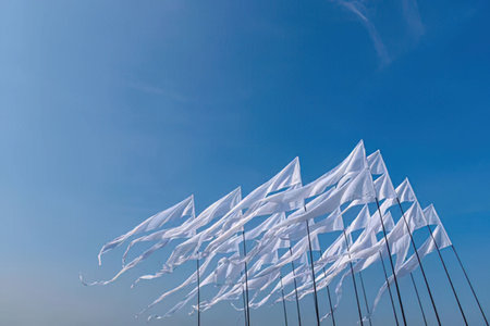 Multiple white flags are waving gently in the wind against a clear blue sky on tall flagpoles...