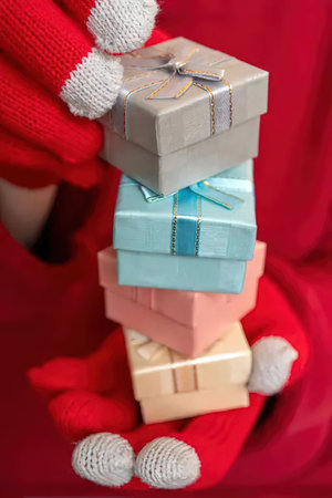 Person wearing red gloves holds a stack of beautifully wrapped gift boxes in various pastel colors...