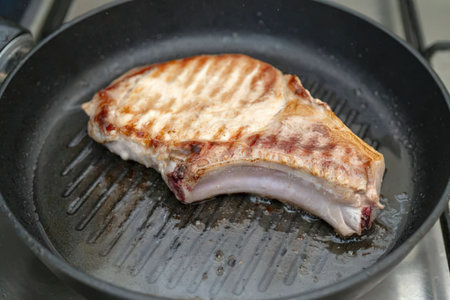 Grilled pork chop cooks in a hot skillet on a stovetop, displaying visible grill marks and a...