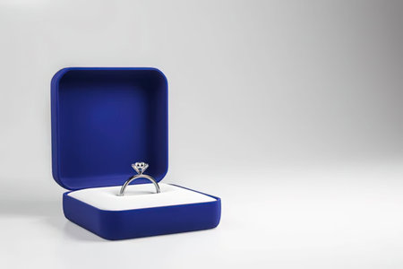 a large diamond ring sitting in a blue jewelry box on a white surface. the ring is sitting on a white interior of the box. the box is open and the ring is sitting on the right side of the box. the background is a plain white wall.