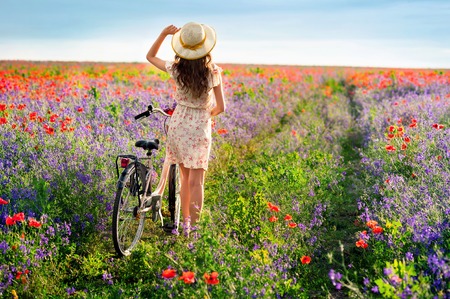 Romantic young woman standing by the back with bicycle on a poppy meadowの写真素材