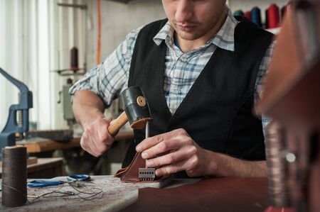 Close up of leather craftsman working with natural leather using hammer. Handbag master at work in...