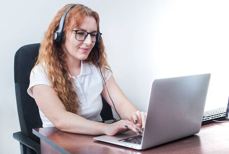 Young beautiful woman with curly hair, wearing glasses, working in the office, uses a laptop and...
