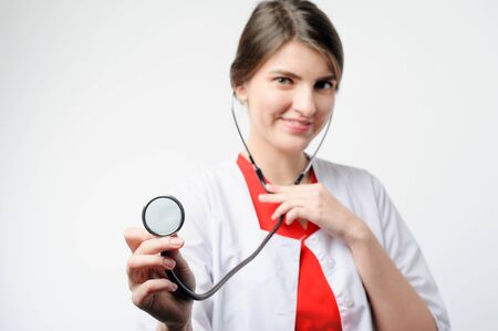 Portrait of smiling lady medical professional in white coat holding phonendoscope looking directly at the cameraの写真素材