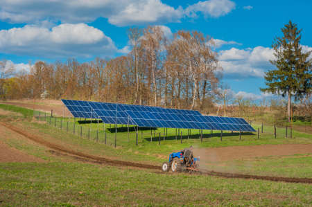 farmers tractor plowing, spring work on field and solar panel with cloudly skyの写真素材
