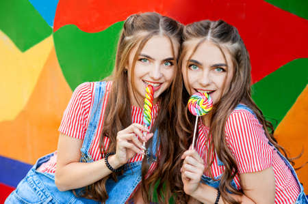 teenage twin sisters in colorful clothes with lollipops on colorful graffiti backgroundの写真素材