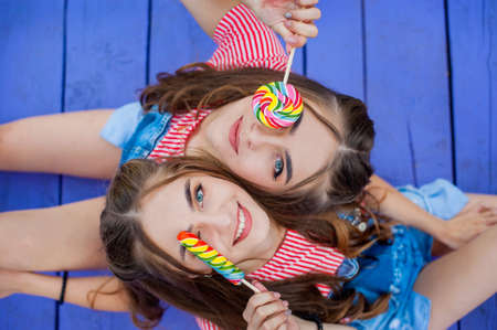 Beautiful teenage twin sisters in colorful clothes with lollipops sitting on colored boards top view