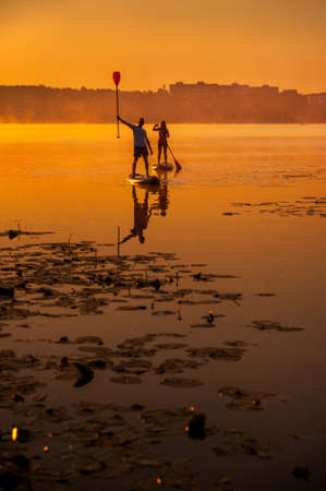 silhouette of a guy with an oar raised above his head, a girl on a sapboard on the water and at sunrise with morning mistsの写真素材