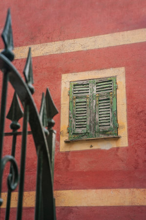 a window with green wooden shutters and a fragment of a wrought iron fence on an ancient red textured facade of an old house Italy, Liguria, Europe.の写真素材