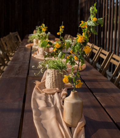 A long wooden table is decorated with yellow flowers in wooden vase, wicker baskets stand on rustic fabric, outdoor celebration.の写真素材