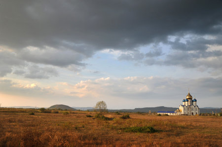 A golden-domed Orthodox church and a tree stand alone in the middle of a wide field under the evening sky, softly illuminating the houses and dry grass, creating a color contrast.の写真素材