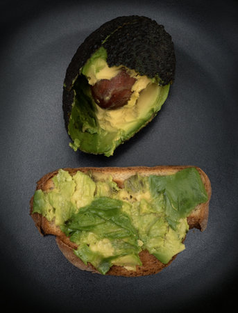 Minimalist composition of a halved avocado and toast with mashed avocado on a dark background. The image highlights natural texture, healthy food, and simplicity in modern culinary aesthetics.の写真素材