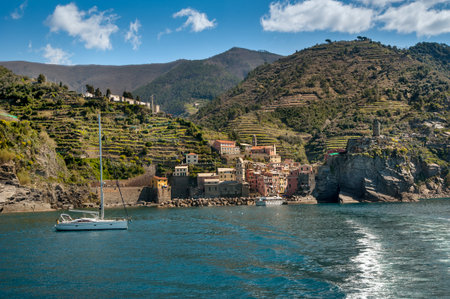 seaside village of Vernazza, sunny coast of Cinque Terre with crystal clear water and green terraces on the slopes. A calm bay with a sailboat near the rocky shore creates an atmosphere of Italian harmony.の写真素材
