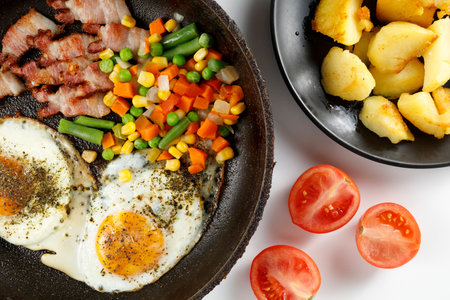Fried eggs with vegetables, fried bacon and potatoes. Hearty food and a delicious lunch.の写真素材