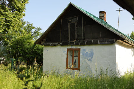 A weathered house with a sloped roof stands in a lush garden under clear skies. The surrounding grass is tall and unkempt.の写真素材
