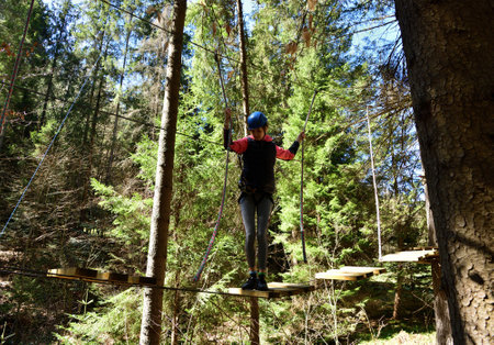 Young woman overcomes wooden obstacles at heights holding on to a rope in amusement parkの写真素材