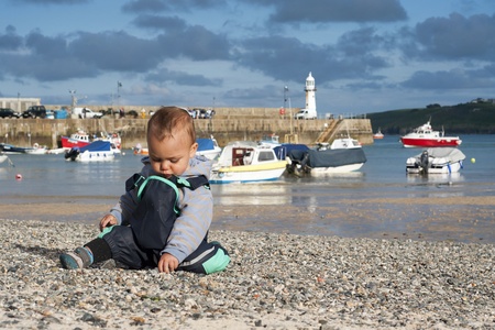 Toddler child, boy, on pebble seaside beach in the village port or harbour; boats and peer in the...