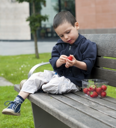 Child eating fresh strawberries from a plastic tub on a bench in a town or city park の写真素材