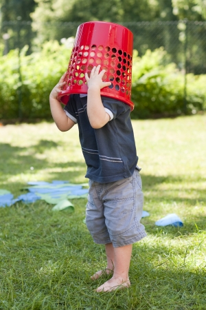 A child toddler with a red basket on his head on a grass in the garden の写真素材