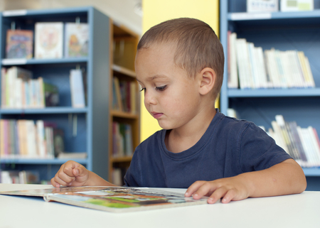 Child boy reading a book in a public library room.