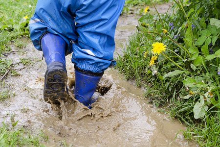 Child in boots walking and jumping in muddy puddle after rain.