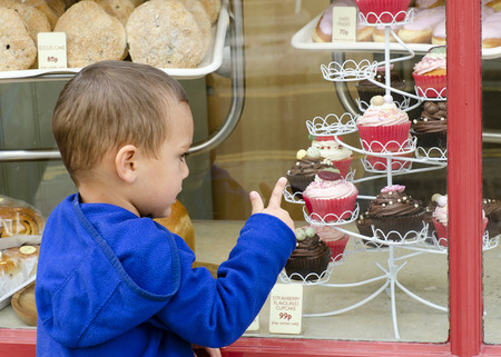 Child looking through the window into sweet cake shop or bakery pointing to cakes.の写真素材