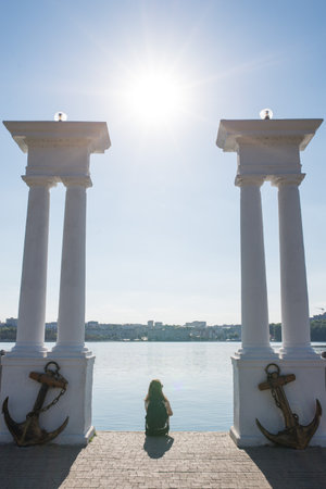 Girl sits on the shore of the lake and looks at the water, on the sides of her two columns with...