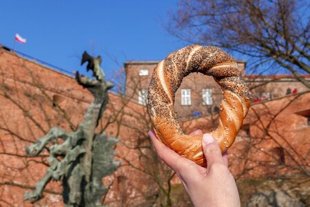 Woman hand holding the Obwarzanek krakowski - the famous Cracow bagel with poppy seed and sesame. The Wawel Dragon in the background. Horizontal imageの写真素材