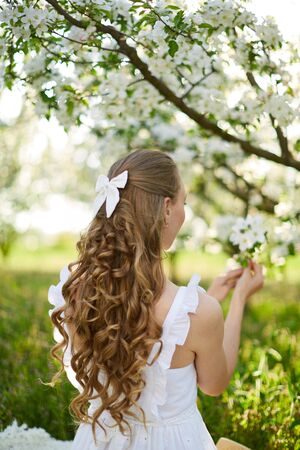 The beautiful girl the blonde in the blossoming apple-trees garden. White flowers in a garden in sunshine. Spring apple trees in blossom. Beautiful woman among spring blossomの写真素材