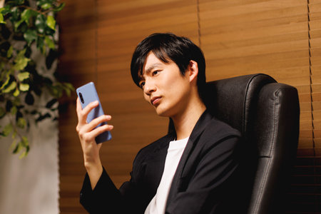 Young Japanese man looking at his phone while sitting in the officeの写真素材