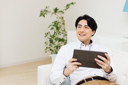 Japanese man smiling and using a digital tablet on sofaの写真素材