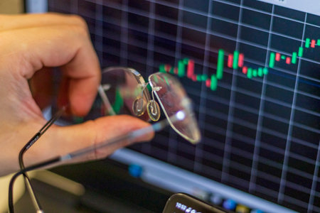 Close up shot of the hand with glasses next to the computer screen with trading bar charts
