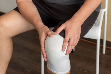 Shot of the man sitting on the white chair, holding his knee with his hands, elastic bandage on the right kneeの写真素材