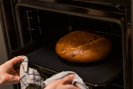 Woman takes out freshly baked loaf of rye bread on the tray with linen cloth from the ovenの写真素材