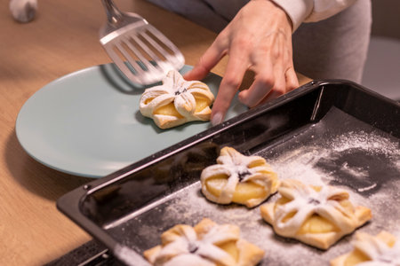 A woman pick the fruit puff pastries with spatula from baking tray and places them on ceramic plateの写真素材