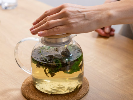 Woman closes wooden lid of the stylish transparent tea pot with just brewed green tea leaves, mint leaves and lemon slices in itの写真素材