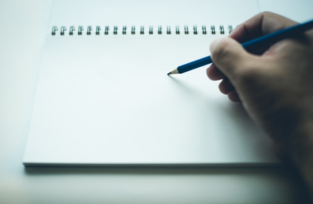 Hand holding a pencil, writing on blank white notebook, with copy space. Shallow depth of field...