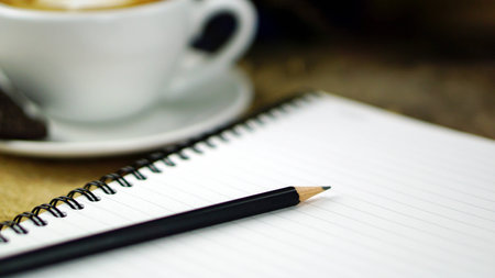 Pencil, memo notebook and cappuccino coffee. A cup of latte, cappuccino or espresso coffee with milk put on a wood table with dark roasting coffee beans.の写真素材