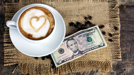 Cappuccino coffee and US Dollars bills. A cup of latte, cappuccino or espresso coffee with milk put on a wood table with dark roasting coffee beans. Drawing the foam milk on top.の写真素材