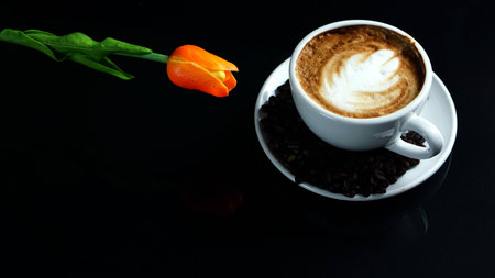 Hot cappuccino with streamed milk. Pour the hot latte art by milk. A cup of strong espresso coffee put on the black glass background with shiny dark roasted Italian coffee beans. Aroma and flavor coffee beverage.の写真素材