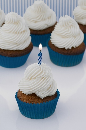 Freshly baked red velvet cupcake with cream cheese frosting and a single burning spiral birthday candle standing in front of several more cupcakesの写真素材