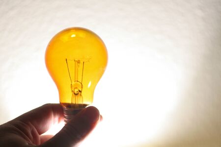 Fingers holding an orange light blub in front of a white wall.の写真素材