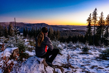 Woman enjoying clear view of winter sunset, sitting on frozen stumpの写真素材