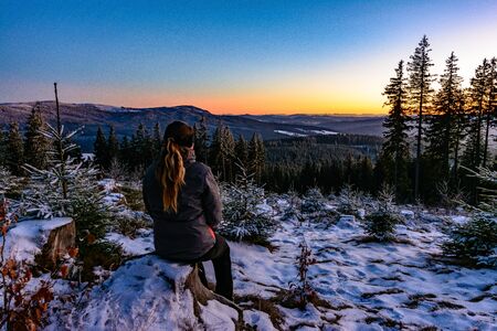 Woman enjoying clear view of winter sunset, sitting on frozen stumpの写真素材